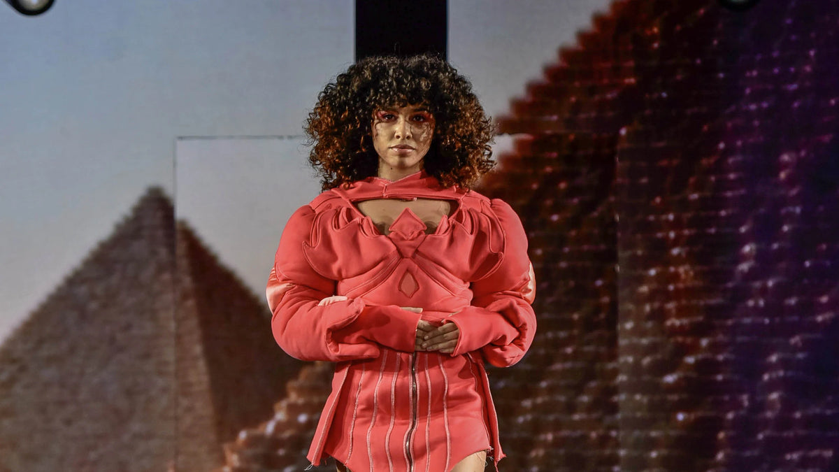 Sculptural coral puffer and pleated skirt walking beneath the projected pyramid
